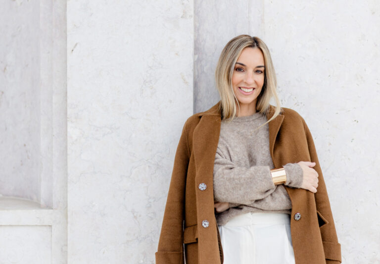 Filipa Simões de Freitas, de casaco camel e camisola bege, encostada a uma parede de pedra clara